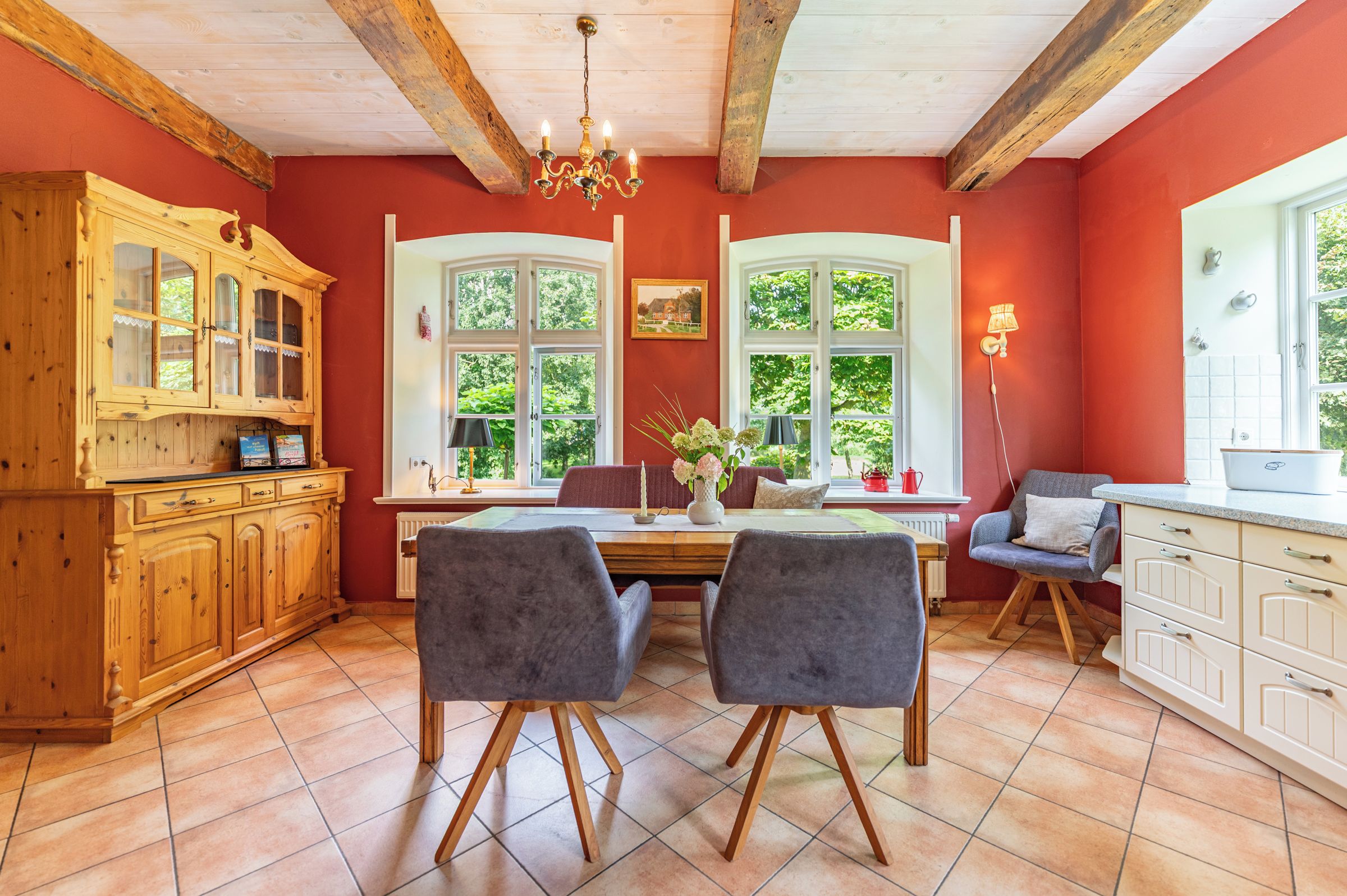 A cozy kitchen with wooden cabinets, a dining table with chairs, and large windows with greenery outside.