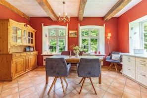 A cozy kitchen with wooden cabinets, a dining table with chairs, and large windows with greenery outside.