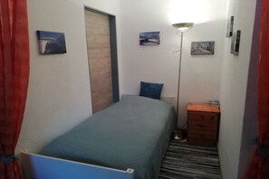 Habitació petita amb un llit individual, una tauleta de nit, una làmpada i un petit calaixer.