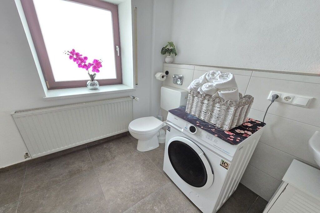 Et lille badeværelse med vaskemaskine, toilet, radiator, et vindue med en plante og en kurv med håndklæder.