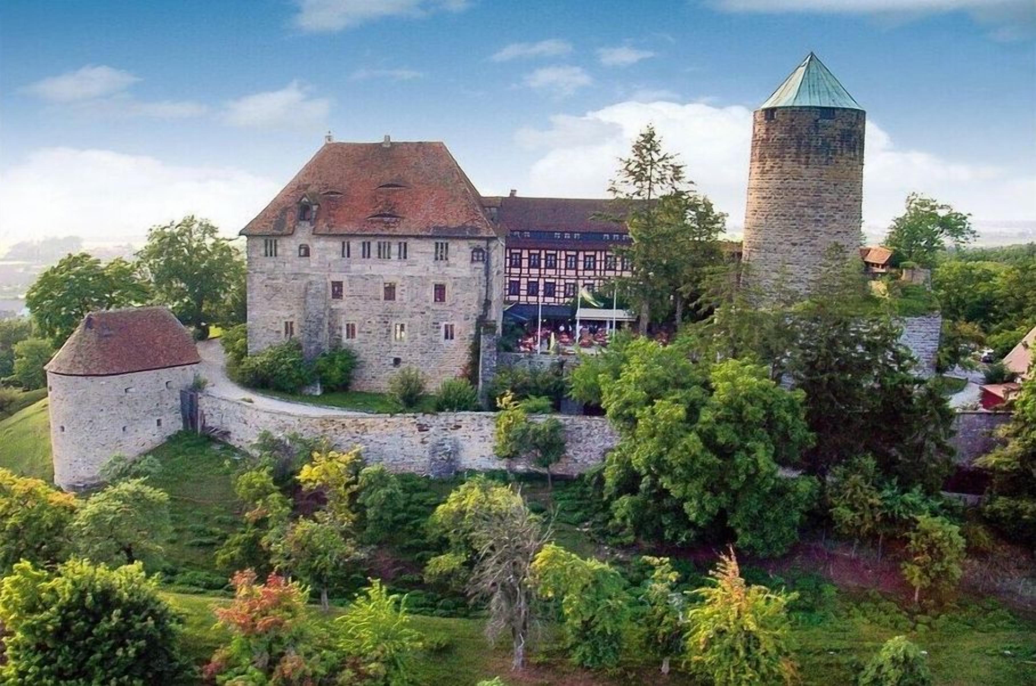 A historic stone building with a tower, surrounded by greenery and a clear sky.