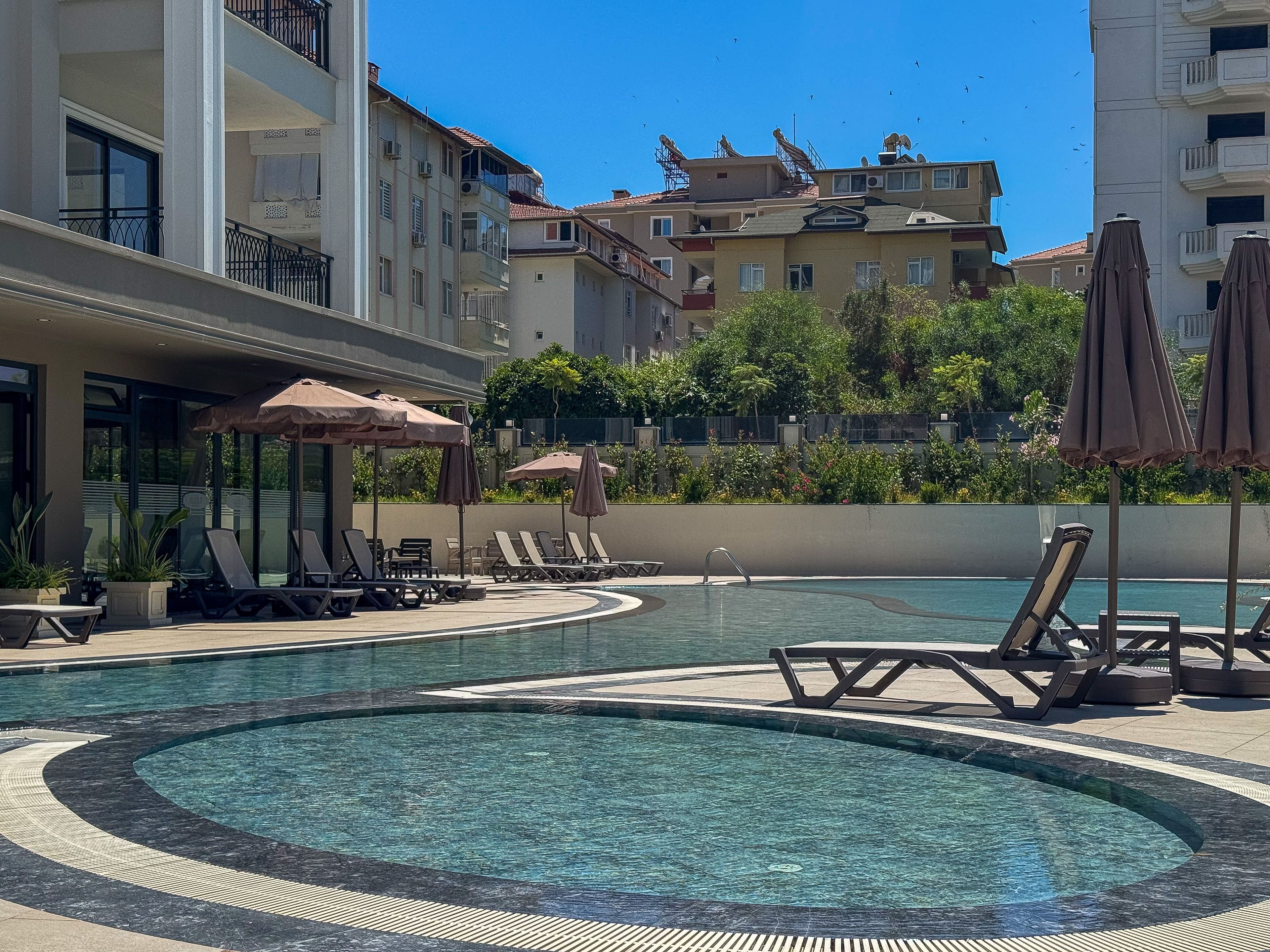 Un espace piscine doté de chaises longues et de parasols, entouré d’immeubles à plusieurs étages.
