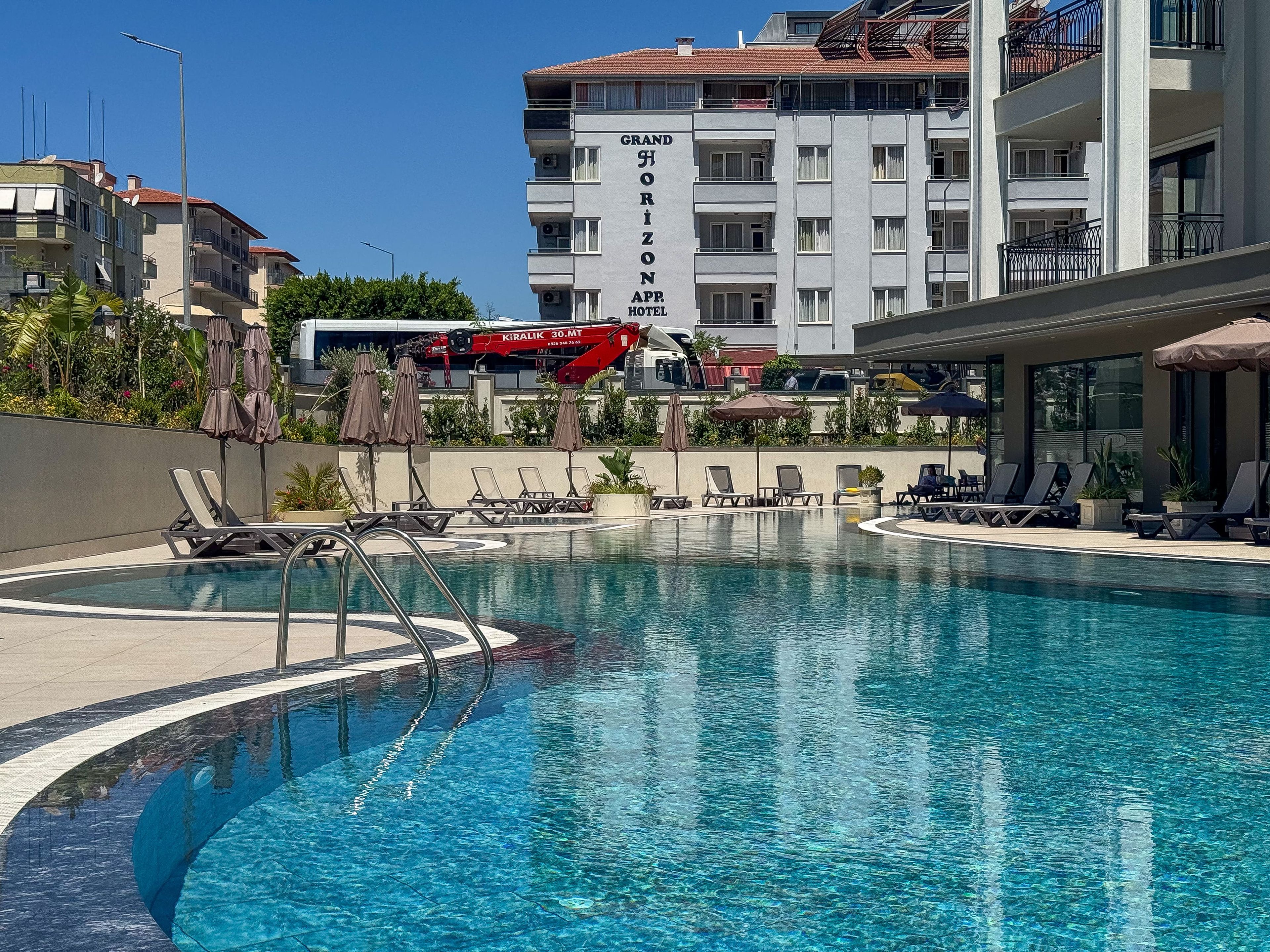A swimming pool with lounge chairs and umbrellas, a red crane, and a building labeled 'Grand Horizon APR Hotel'.