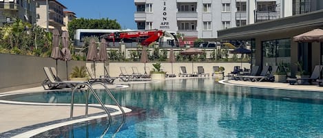 A swimming pool with lounge chairs and umbrellas, a red crane, and a building labeled 'Grand Horizon APR Hotel'.