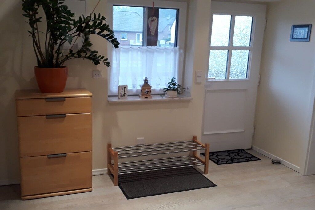Una habitación con un aparador de madera, una planta en maceta, una ventana con cortina, un estante pequeño con adornos y un felpudo en el suelo.