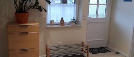 Una habitación con un aparador de madera, una planta en maceta, una ventana con cortina, un estante pequeño con adornos y un felpudo en el suelo.