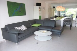 Ein modernes Wohnzimmer mit einem grauen Ecksofa, einem runden Couchtisch und einem Essbereich mit gemusterten Stühlen.