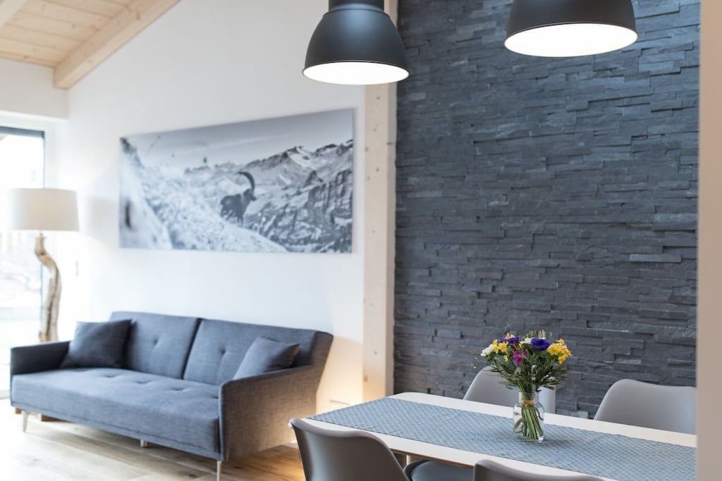 A modern living room with a grey sofa, a dining table with chairs, a vase of flowers, and a black pendant light.