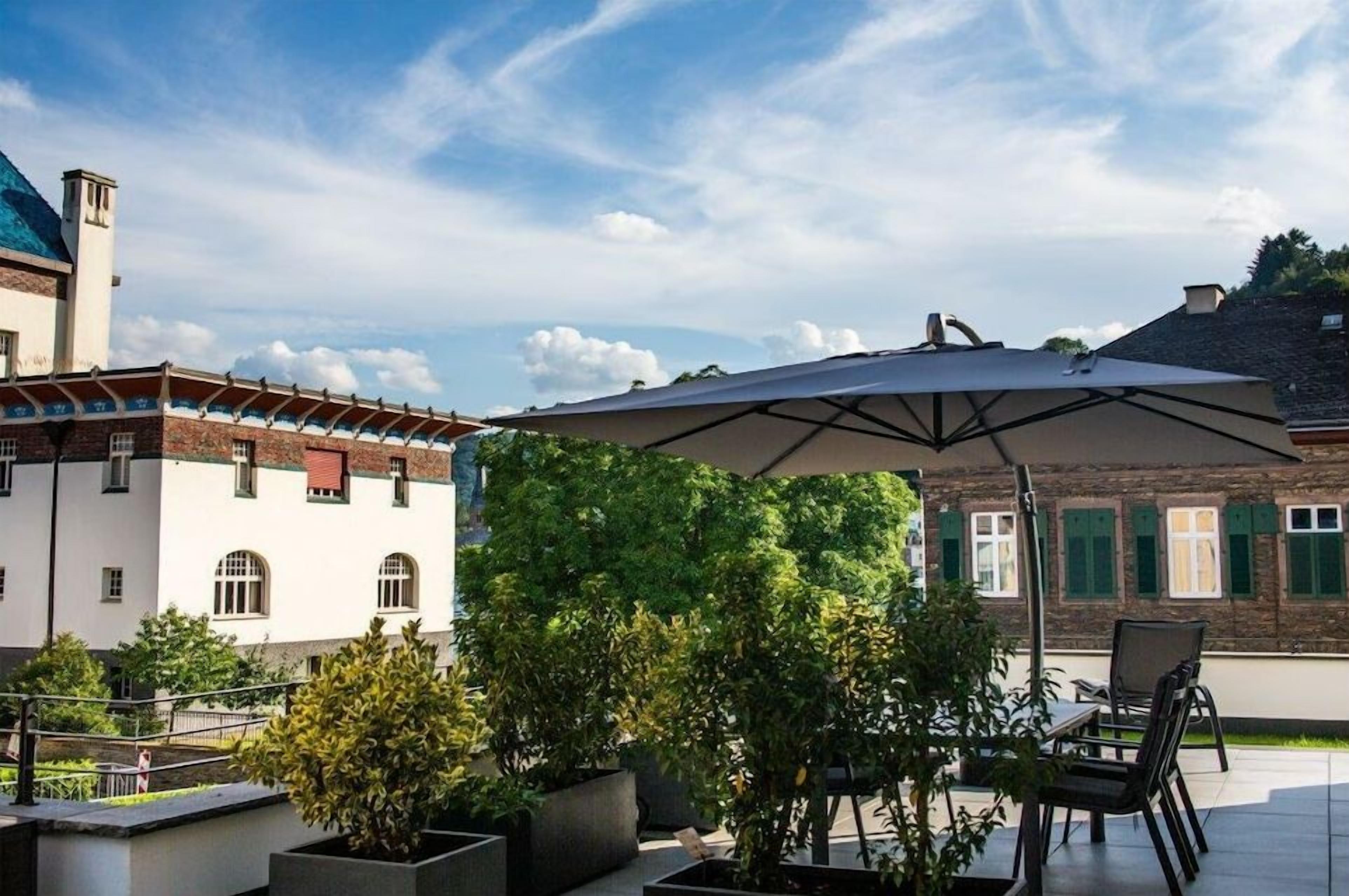 En udendørs patio med et bord og stole under en stor parasol, omgivet af krukker med planter og en bygning med murstensfacade.
