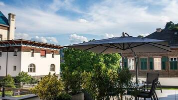 En udendørs patio med et bord og stole under en stor parasol, omgivet af krukker med planter og en bygning med murstensfacade.