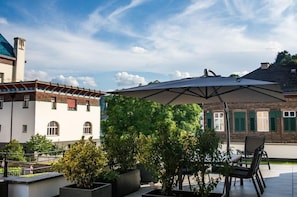 En udendørs patio med et bord og stole under en stor parasol, omgivet af krukker med planter og en bygning med murstensfacade.
