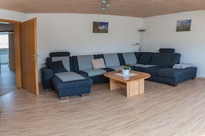 Uma sala de estar moderna com um sofá seccional, uma mesa de centro de madeira e uma porta que dá para outro cômodo.