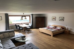 Une chambre à coucher avec un lit en bois, un canapé gris, une table basse en verre et un coin repas comprenant des chaises et une table.