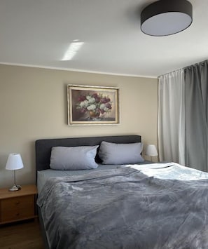 A bedroom with a bed, two bedside lamps, a nightstand, a framed floral artwork on the wall, and a window with curtains. - Holiday apartment with 1 bedroom (Schönau am Königssee)