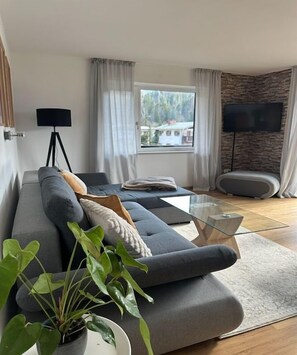 Uma sala de estar moderna com um sofá, uma mesa de centro de vidro, um vaso de planta, uma televisão e uma janela com cortinas.