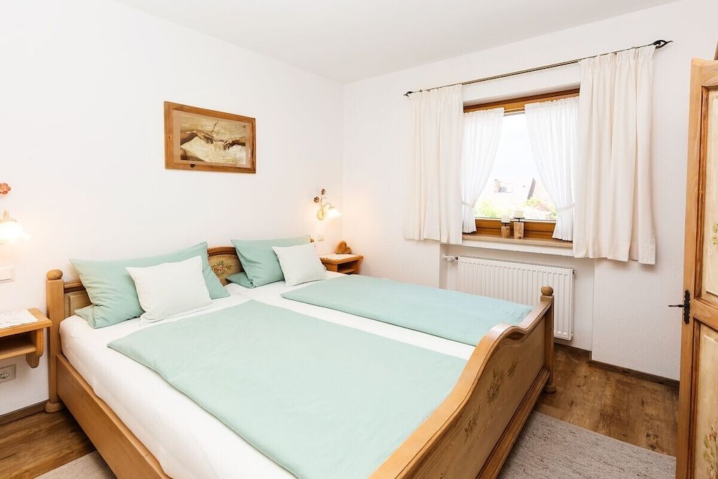 Una cama doble con estructura de madera, dos mesitas de noche, una lámpara de noche, una ventana con cortinas, un radiador y un cuadro colgado en la pared.