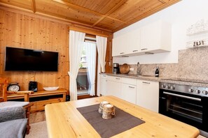 Une pièce aux murs lambrissés, équipée d’un téléviseur, d’une table à manger avec un set de table, et d’une cuisine avec des meubles blancs et une cuisinière.