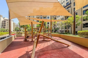 An outdoor playground with wooden equipment and a red rubberized surface, surrounded by greenery and modern buildings. - Resort style 2 bedroom by Jumeirah Beach (Dubai)