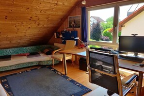 Ein gemütliches Zimmer mit schrägen Wänden, einem Bett mit einem grün gemusterten Überwurf, einem Schreibtisch mit einem Computer und einem Fenster mit Blick auf Grünflächen.