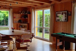 A room with a pool table, a dining table with chairs, and a large window with a view of greenery. - Wooden house with large garden (Uhldingen-Mühlhofen)