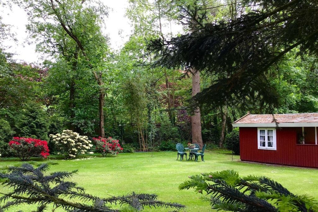 A red building with a white window, surrounded by a well-maintained garden with various plants and a lawn.