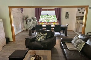 Een woonkamer met een bank, een bijzettafeltje met decoratieve objecten, een eethoek met een tafel en stoelen, en uitzicht op groen buiten.