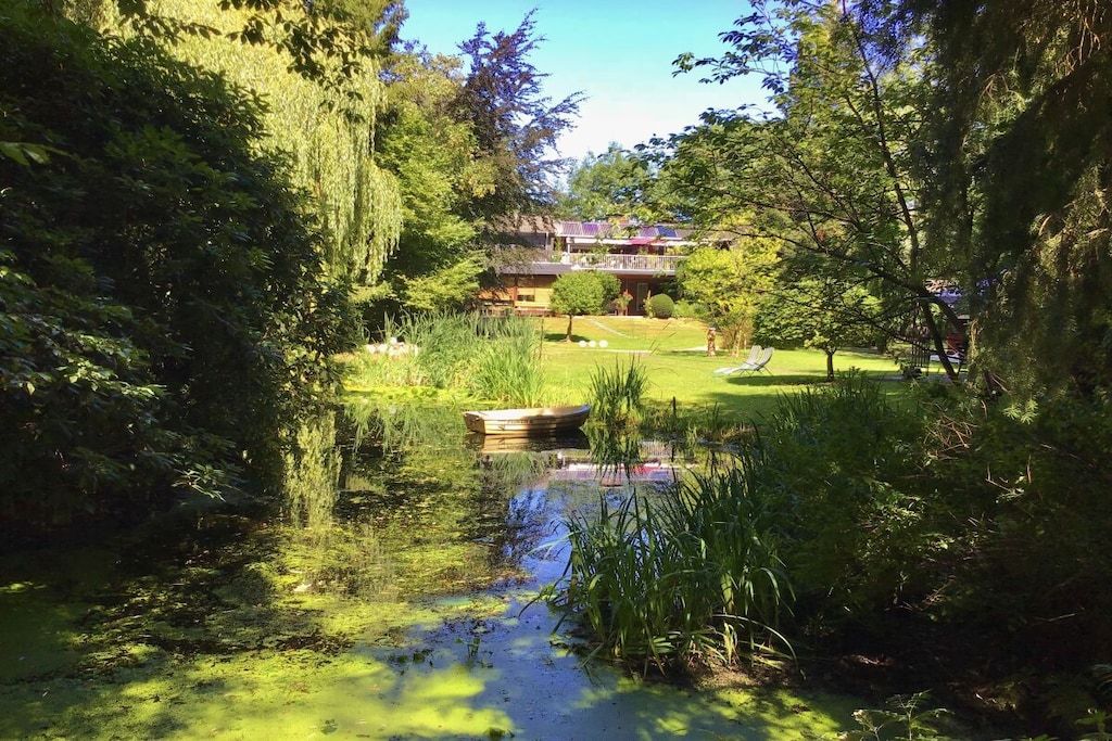 Un jardín tranquilo con un estanque, abundante follaje y un edificio con balcón al fondo.
