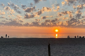Strandszene mit Sonnenuntergang, Silhouetten von Personen und einem Hund, und einem Holzpfosten im Vordergrund.