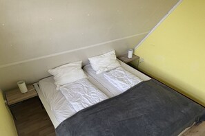 Ein kleines Zimmer mit einem Einzelbett, Holzboden, grauer Bettdecke und weißer Wand.