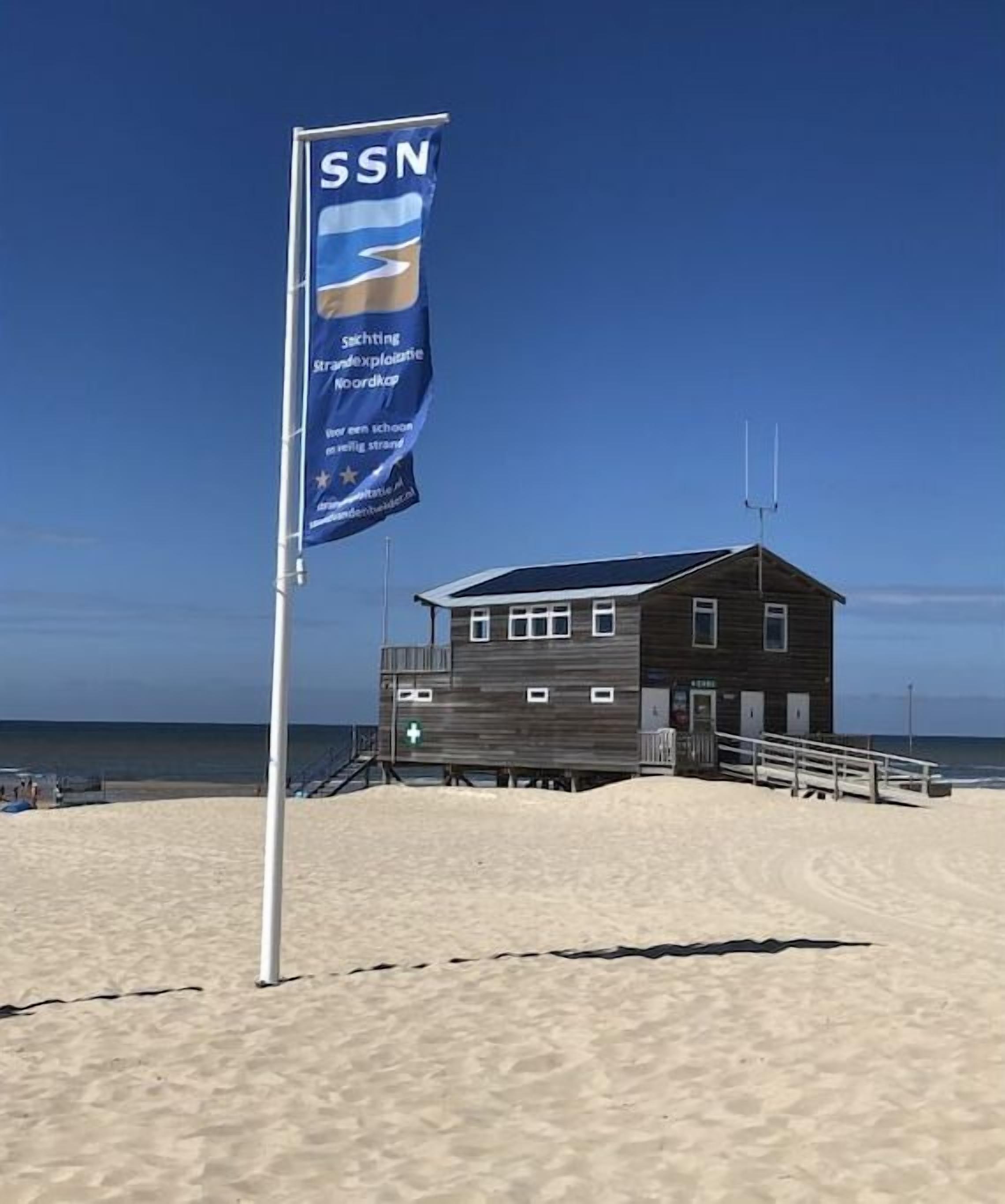 Un bâtiment situé au bord de la plage, avec un drapeau, une tour de surveillance pour les sauveteurs et un ciel dégagé.