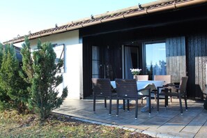 An outdoor patio with a table and chairs, a small tree, and a building with a tiled roof. - Holiday home "Breitenberg" (Lechbruck)