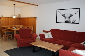 A living room with a red sofa, a wooden coffee table, and a dining area with a wooden table and chairs. - Holiday home "Breitenberg" (Lechbruck)