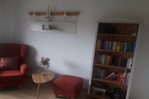 A living room with a red armchair, a small wooden table with a vase, a red ottoman, and a bookshelf filled with books. - Holiday apartment with 1 bedroom (Wurster Nordseeküste)