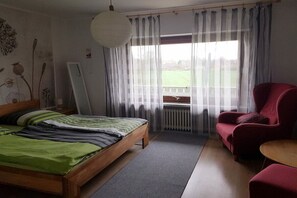 Ein Schlafzimmer mit einem Bett, einem roten Sessel, einem kleinen Tisch und einem Fenster mit Vorhängen.