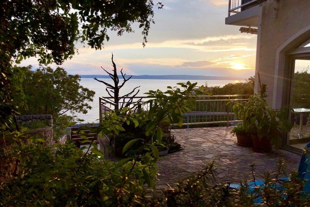 Un patio de piedra con plantas en macetas, una verja de metal y vista al mar al atardecer.