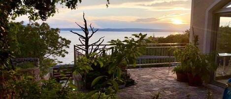 Un patio de piedra con plantas en macetas, una verja de metal y vista al mar al atardecer.