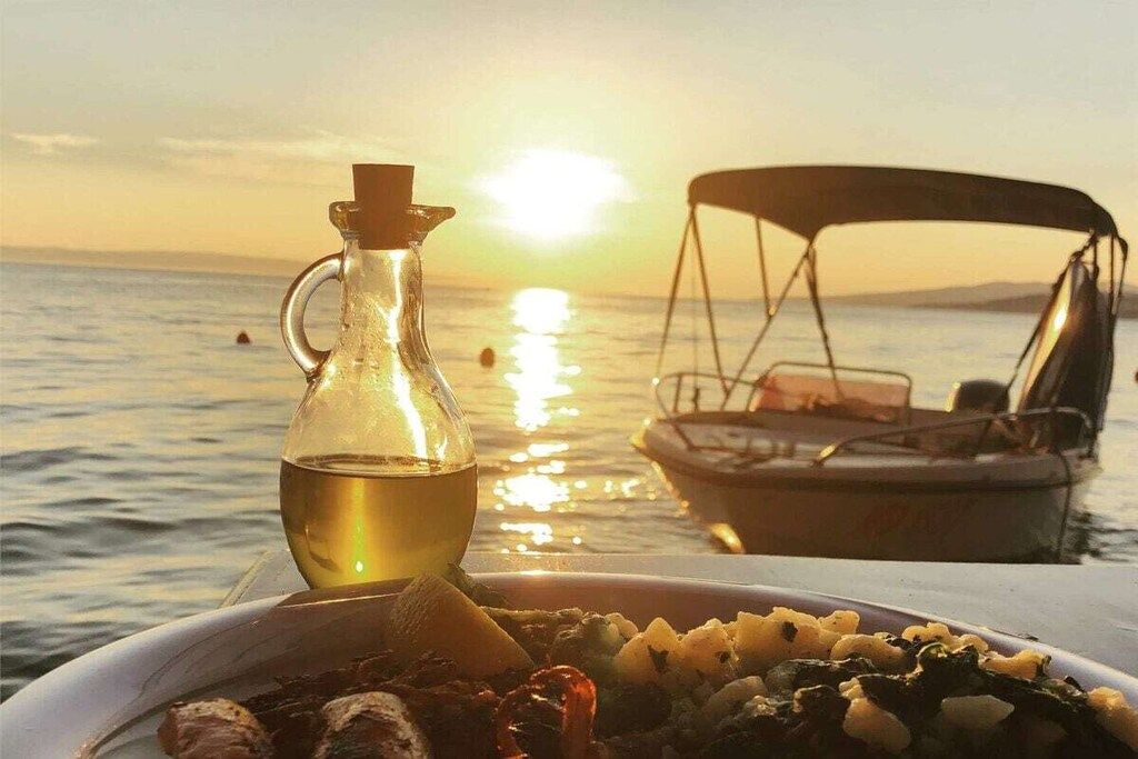 Une table avec une assiette de nourriture et une bouteille d’huile, un bateau en arrière-plan et un coucher de soleil sur l’eau.