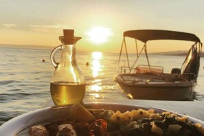 Ein Tisch mit einer Speiseplatte und einer Ölflasche, ein Boot im Hintergrund und ein Sonnenuntergang über dem Wasser.