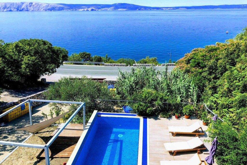 Ein Schwimmbecken mit Blick auf das Meer, umgeben von Grünpflanzen und einer Straße.
