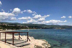 Eine Holzterrasse mit Blick auf eine Küstenlandschaft, klares blaues Wasser und in der Ferne Hügel.
