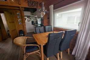 A dining area with a wooden table and chairs, a wicker chair, a hanging light fixture, and a view through a window with curtains. - with garden directly on the water (wooden house) (Lemmer)