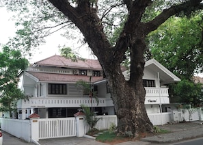 A two-story building with a tiled roof and a white facade, surrounded by trees and a white fence.