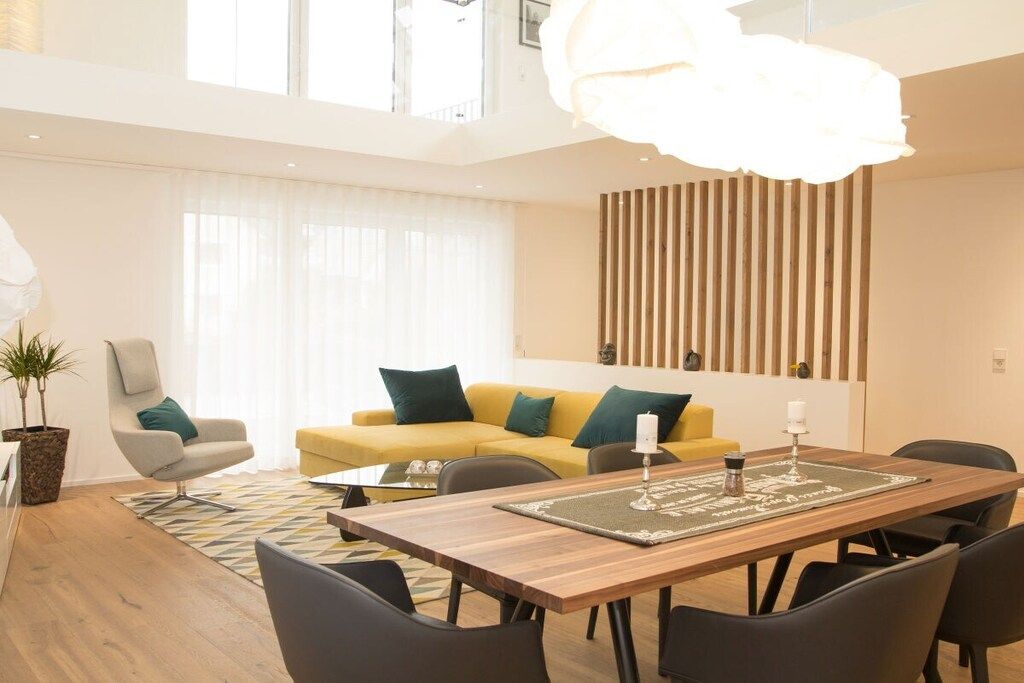 A modern living room with a wooden dining table, black chairs, a yellow sofa with green cushions, a white armchair, and a large window with sheer curtains.