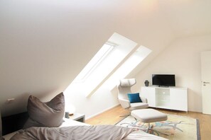 Ein modernes Schlafzimmer im Loft-Stil mit schrägverdeckter Decke, einem Bett, einer Sitzecke mit Sessel und Hocker, einem Fernseher auf einem weißen Schrank und einem großen Fenster, das für natürliches Licht sorgt.
