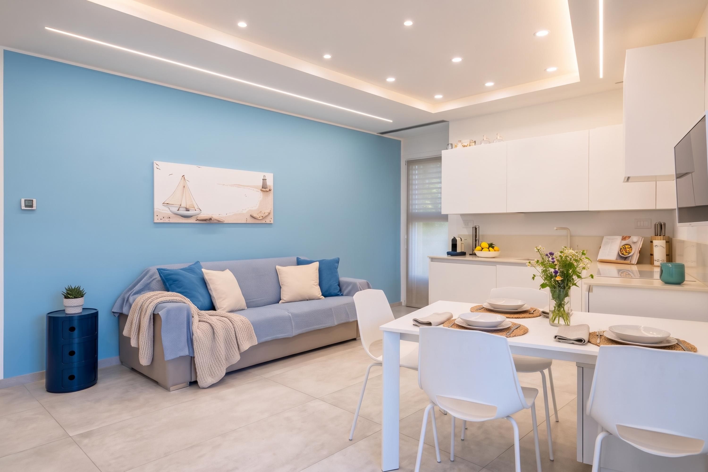 Een moderne woonkamer met een bank, een eettafel met stoelen en een keukenhoek met kasten en een raam.