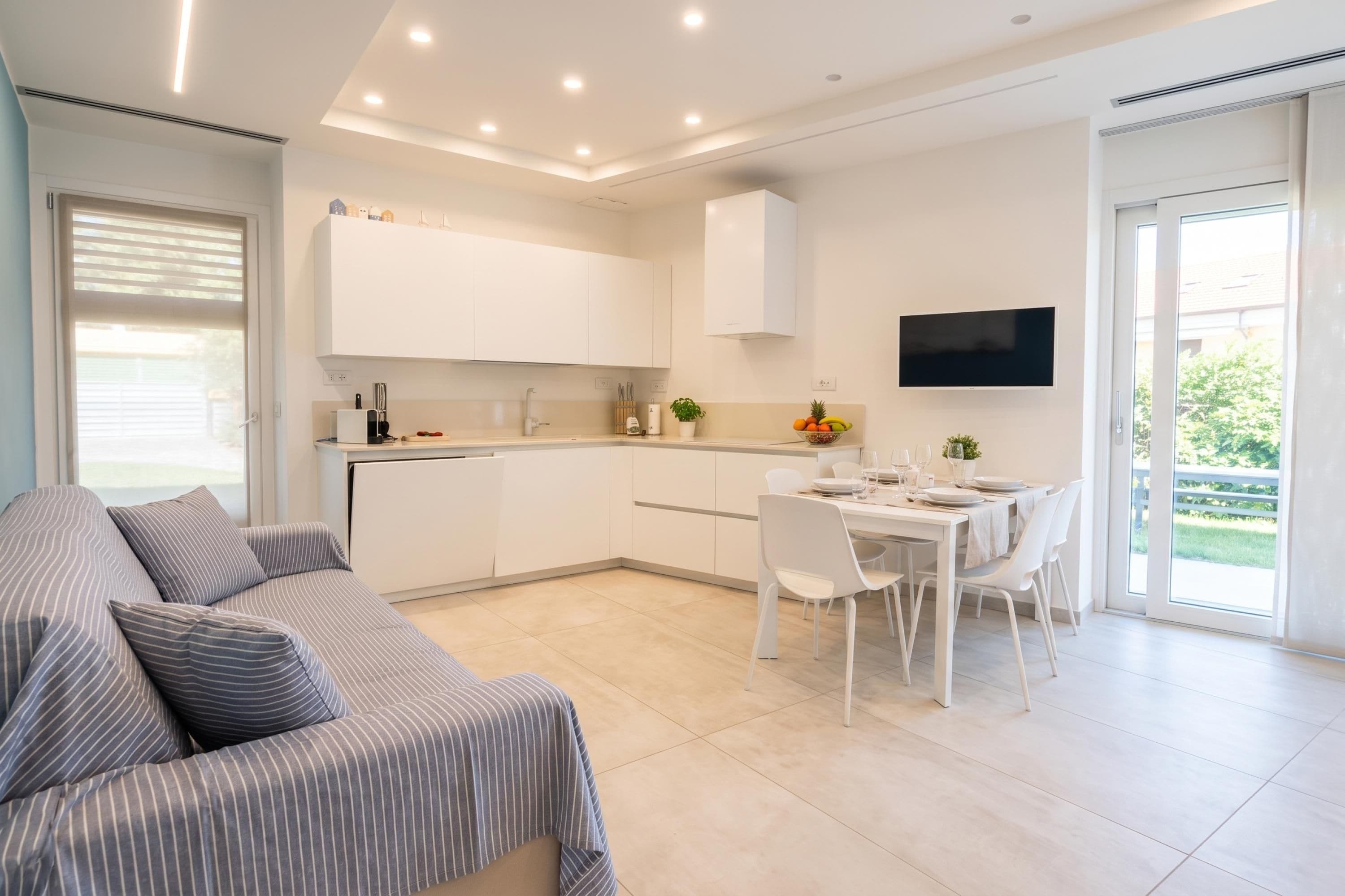 A modern kitchen with white cabinets, a dining area with a table set for four, and a large window providing a view of the outdoors.