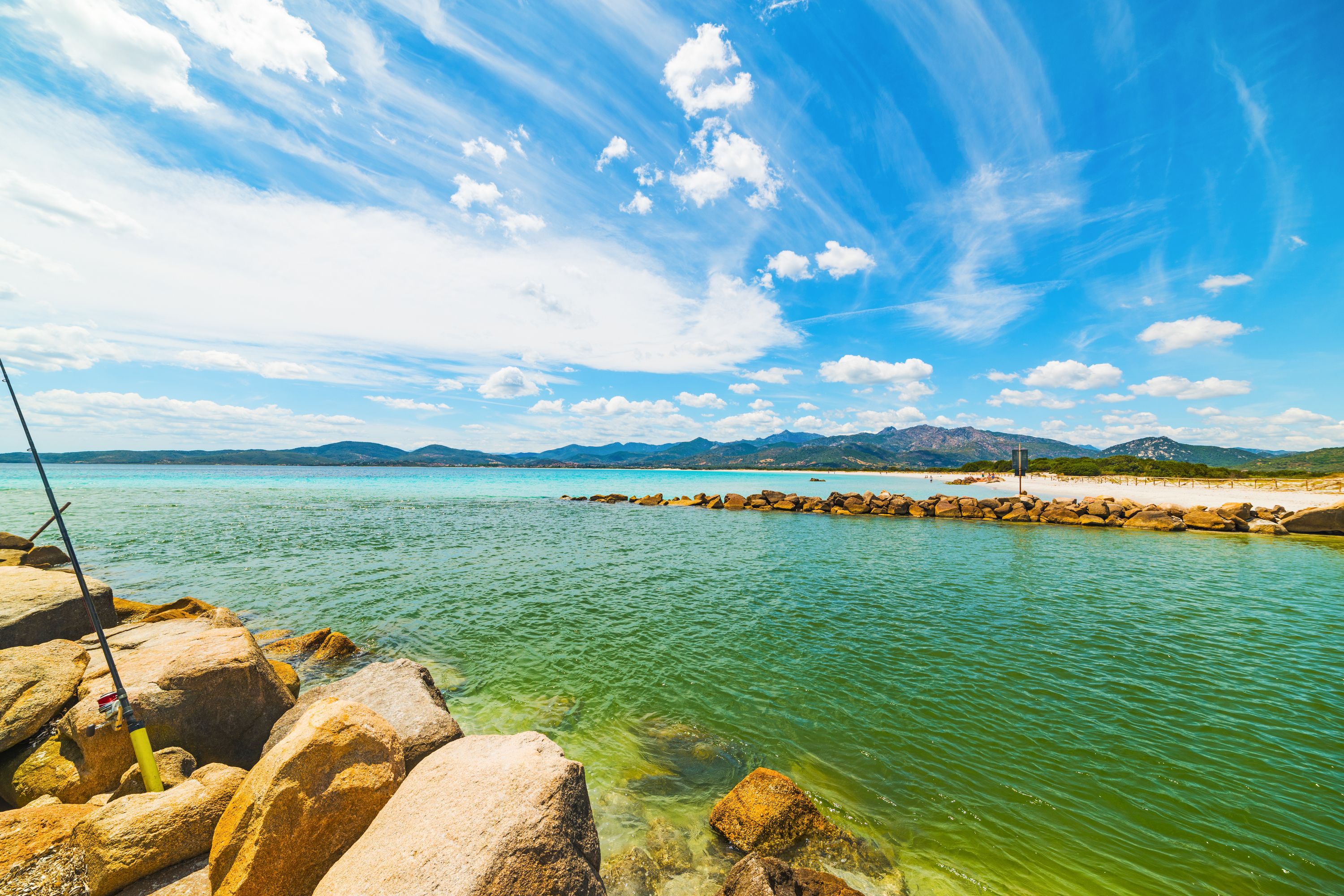 A rocky shoreline with clear turquoise water, a fishing rod, and distant mountains under a blue sky.