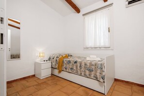 A bedroom with a bed, a bedside table with a lamp, a window with curtains, and a white wall. - Casa Molara (San Teodoro)