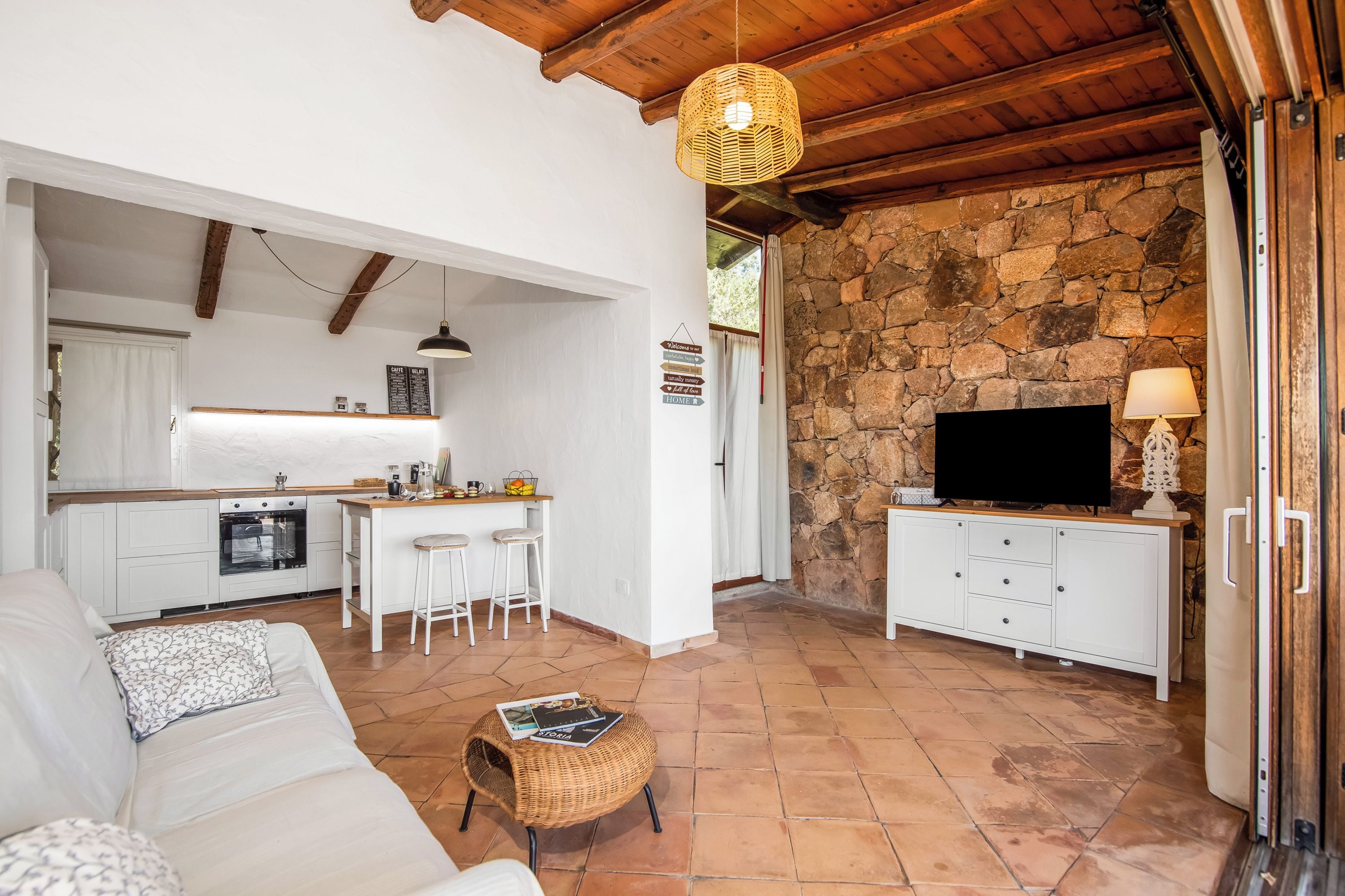 A modern living room with a sofa, a wicker ottoman, and a kitchen area with a stone wall and a television.