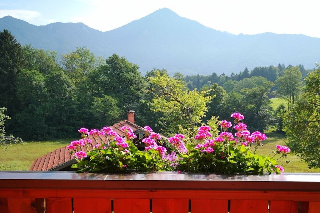 Un jardin aux fleurs roses devant une maison avec un toit rouge, entouré d’arbres verdoyants et avec des montagnes en arrière-plan.